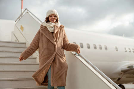 Cheerful woman in fur coat walking down airplane stairsの写真素材
