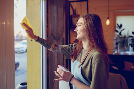 Cheerful young woman cleaning window on cafeteriaの写真素材