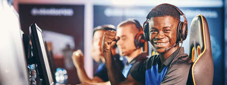Side view of a cheerful african guy, professional cybersport gamer wearing headphones looking at camera and smiling while participating with team in eSport tournamentの写真素材