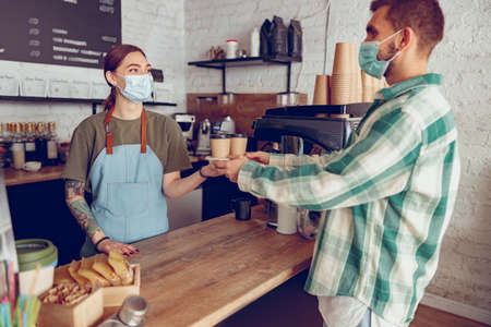 Woman barista in medical mask giving coffee to customer in cafeの写真素材