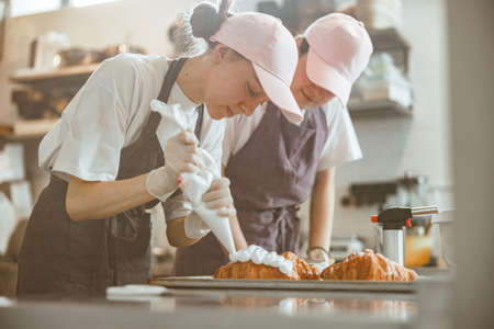 Confectioner trainee applies cream onto croissant under skilled colleague control in bakery shopの写真素材