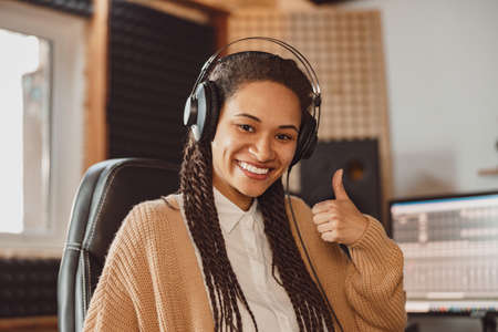 Beautiful African woman musician in headphones shows thumb up, smiles toothy smile looking at camera in recording studioの写真素材