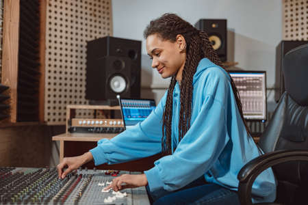Stylish female audio engineer, producer works in music recording studio, uses mixing board, software...
