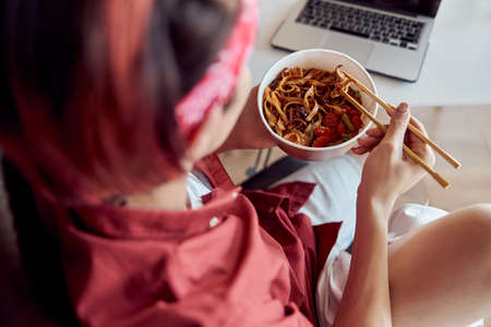 Young man holding chopsticks eating takeaway noodle box meal