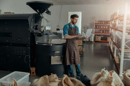 Business owner of coffee factory working laptop on background of coffee roasting machineの写真素材