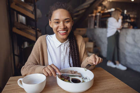 Portrait of cheerful African young woman smiling toothy smile to camera while snacking at a food and drink establishmentの写真素材