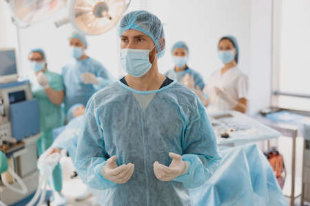 Male surgeon in mask and gloves standing in operating room ready to work on patient and looking away