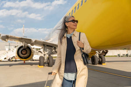 Passenger woman in sunglasses with luggage is outdoors near plane looking awayの写真素材