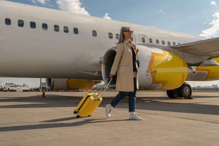 Passenger woman in sunglasses with luggage is outdoors near plane at airport looking away