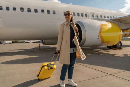Passenger woman in sunglasses with luggage is outdoors near plane at airport looking away