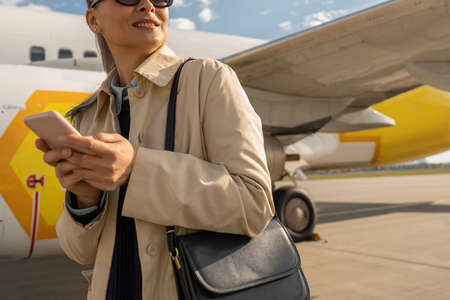 Close up of woman passenger in sunglasses using phone standing outdoors at airport near plane
