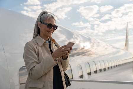Cheerful woman passenger using phone standing outdoors at airport near planeの写真素材