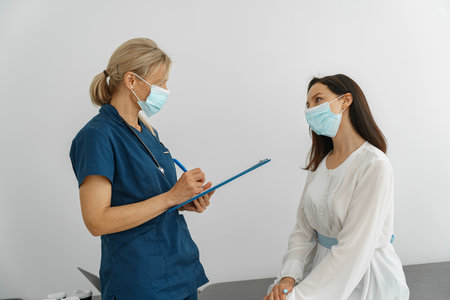 Doctor wearing protection mask against covid taking notes during consultation with patientの写真素材