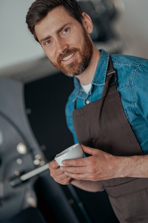Portrait of male barista with cup of fresh coffee on background of roasting machineの写真素材
