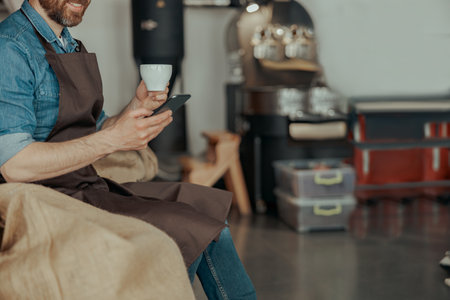Close up of barista with cup of coffee and phone sit on warehouse of coffee factoryの写真素材