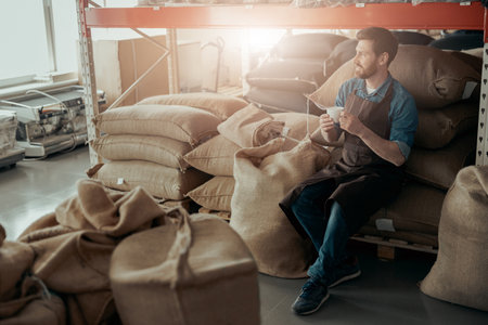 Business owner with cup of coffee and phone sit on warehouse of coffee factory and looking awayの写真素材