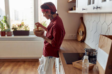 Asian guy standing in the kitchen and eating noodles with chopsticksの写真素材