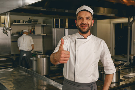 Chef in uniform standing on kitchen of restaurant with colleague on background and showing thumb upの写真素材