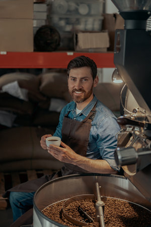 Smiling business owner with cup of fresh coffee on background of roasting machine
