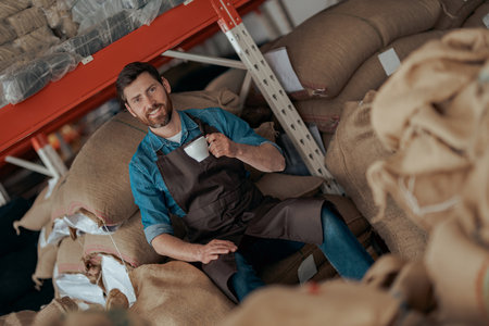 Smiling Business owner with cup of fresh coffee sitting on bags with roasted coffee beansの写真素材
