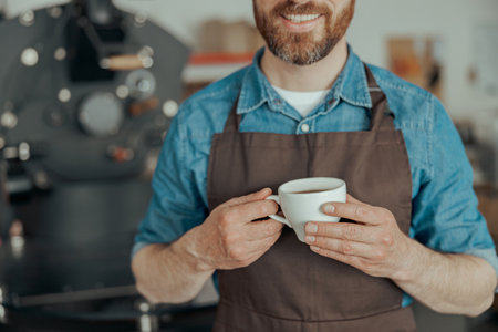 Close up of smiling business owner with cup of fresh coffee on background of roasting machineの写真素材
