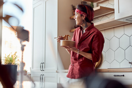 Smiling Asian guy leaning on the kitchen countertop and eating at homeの写真素材