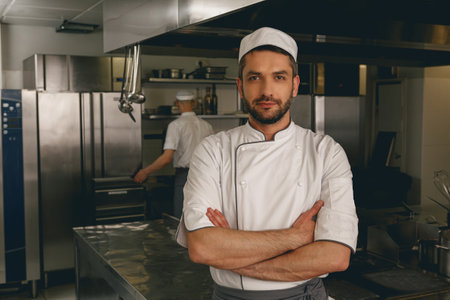 Portrait of male chef in uniform standing on kitchen of restaurant with colleague on backgroundの写真素材