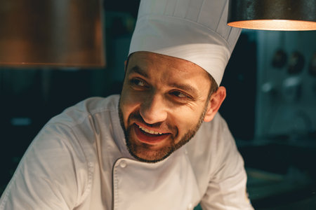 Close up of smiling male chef in uniform standing on kitchen of restaurant and looking awayの写真素材