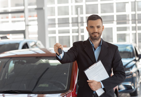Young male consultant showing new car and holding keys to it in dealershipの写真素材