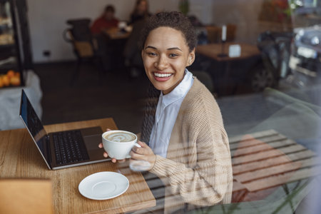 Attractive African American young woman holding a cup with cappuccino, enjoying remote work on laptop in coffee shopの写真素材