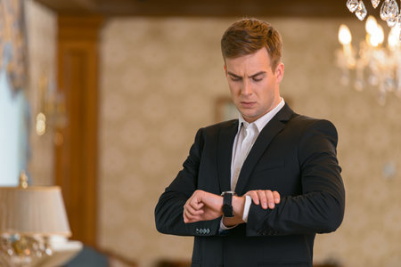 Serious businessman in suit standing in hotel room and looks at the wristwatchの写真素材