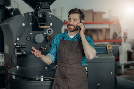 Business owner of coffee factory talking phone on background of coffee roasting machineの写真素材