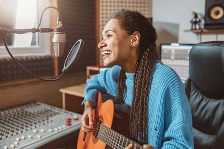 Pleasant cute multiethnic woman singing into microphone and playing guitar in the broadcasting of sound recording studioの写真素材