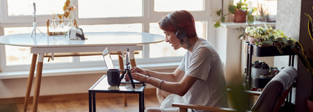 Smiling Asian guy in headphones working with laptop at home while typingの写真素材
