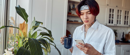 Asian guy typing on mobile phone while holding cup of coffeeの写真素材