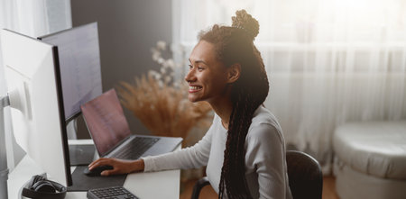 Smiled cheerful female web expert sitting at desk in living room coding on computerの写真素材