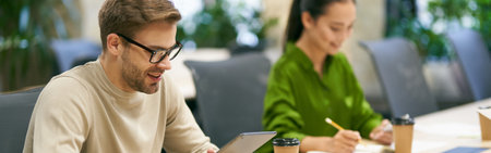 Working together. Young smiling caucasian man wearing eyeglasses using digital tablet while sitting at desk with his female colleague in the modern coworking spaceの写真素材