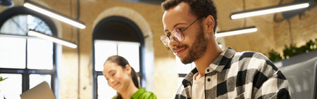 Young smart guy wearing eyeglasses working on laptop while sitting at desk with his female colleague in the modern coworking space or officeの写真素材