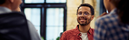 Young smiling mixed race male office worker sitting at the desk and talking with colleagues, working together in the modern officeの写真素材