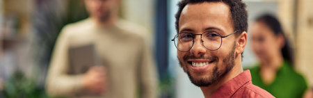 Young cheerful mixed race man holding digital tablet and smiling at camera while standing in the modern office with his colleagues on the backgroundの写真素材