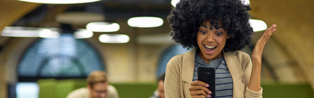 Good news. Young excited mixed race woman using smartphone and smiling while standing in the coworking space with her colleagues working on the backgroundの写真素材