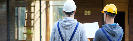 Plan it. Rear view of two builders standing outdoors with an open blueprint discussing the interior of a half completed cottage houseの写真素材