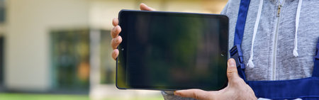 Cropped shot of bearded male builder smiling at camera, holding digital tablet pc with blank screen while working at cottage construction siteの写真素材