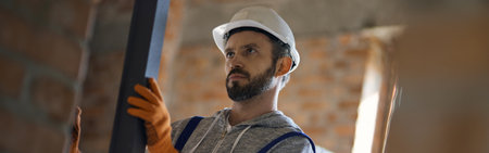 Focused young male builder wearing hard hat holding metal stud for drywall while working at construction siteの写真素材
