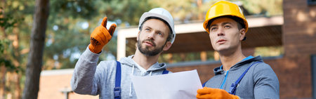 Building ideas. Portrait of two builders looking away while standing outdoors with an open blueprint discussing the half completed cottage houseの写真素材