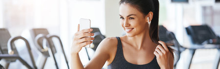 Always beautiful. Happy young woman in sportswear is looking at smartphone screen and smiling while standing at gymの写真素材