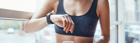Making training easier. Young and cheerful woman in sportwear looking at her sport bracelet and using smartphone while standing in front of window at gymの写真素材