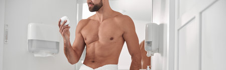 Seductive man holds jar of cream leaning onto cabinet near mirrorの写真素材