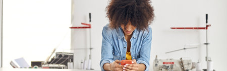 Young African-American woman surfs internet with phone at cutting table in sewing workshopの写真素材