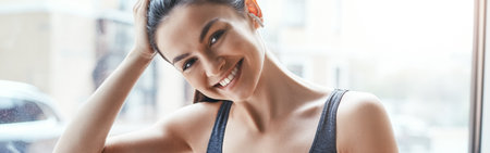 Simply happy. Beautiful and cheerful woman in sportswear and headphones listening music and looking at camera while standing in front of window at gymの写真素材
